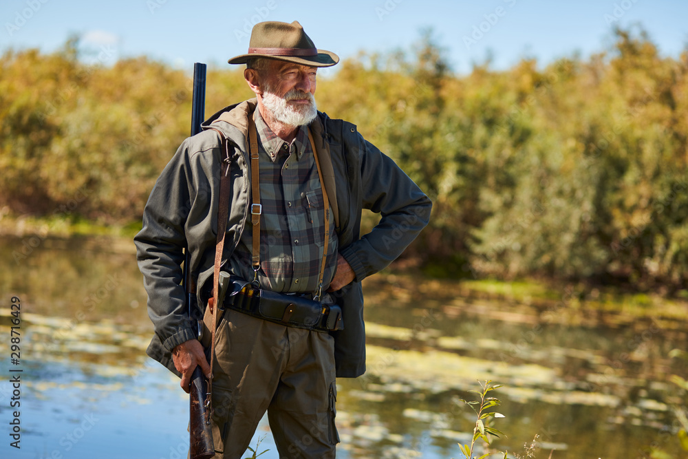 Fototapeta premium Senior man with gun going to hunt on wild ducks on lake. Male looking away, one arm on lower back