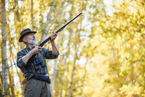 Hunting on wild birds in forest. Caucasian senior man hunt on birds, using gun. Hunter wearing cowboy hat and casual hunting clothes. Forest background