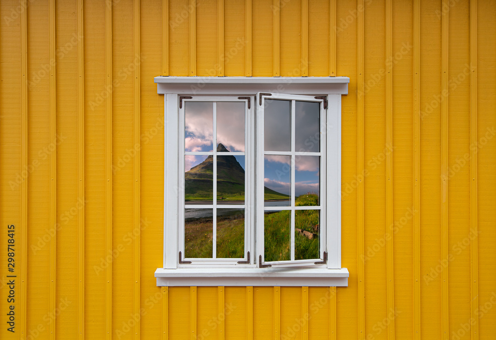 Yellow wooden house with a white window with a reflection of the Icelandic landscape