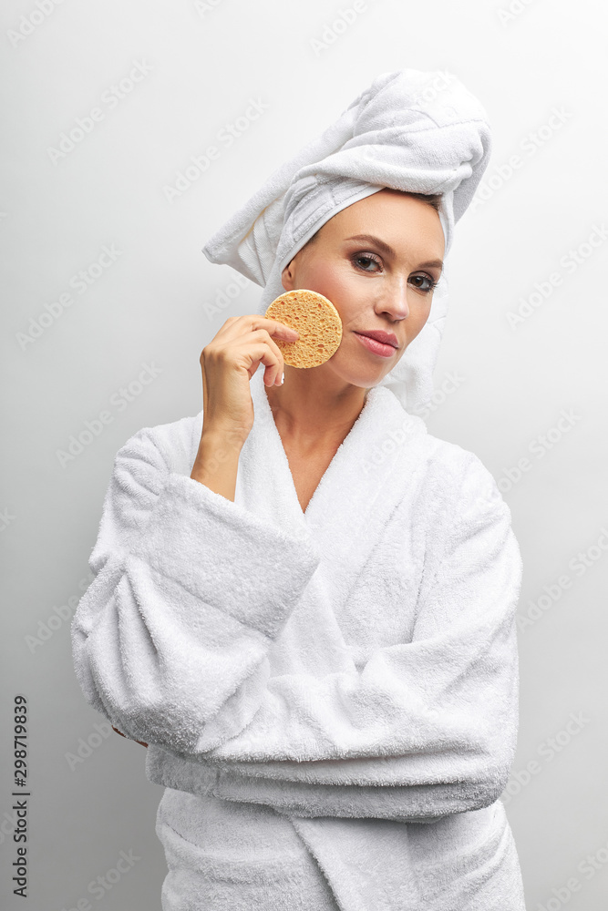 Beautiful lady in bathrobe with white towel on her head and beige rough sponge for scrubbing in her right hand. White isolated background Beautiful lady in bathrobe with white towel on her head and beige rough sponge for scrubbing in her right hand. White isolated background