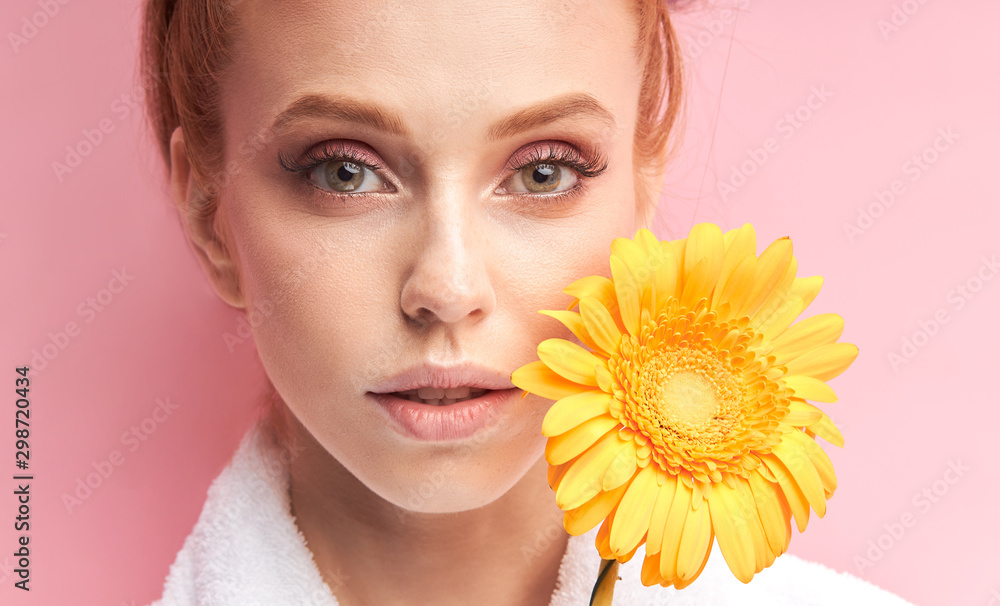 Young caucasian woman holding daisy yellow flower isolated over pink background. Female wearing white bathrobe, curlers on hair after shower. Natural beauty Young caucasian woman holding daisy yellow flower isolated over pink background. Female wearing white bathrobe, curlers on hair after shower. Natural beauty