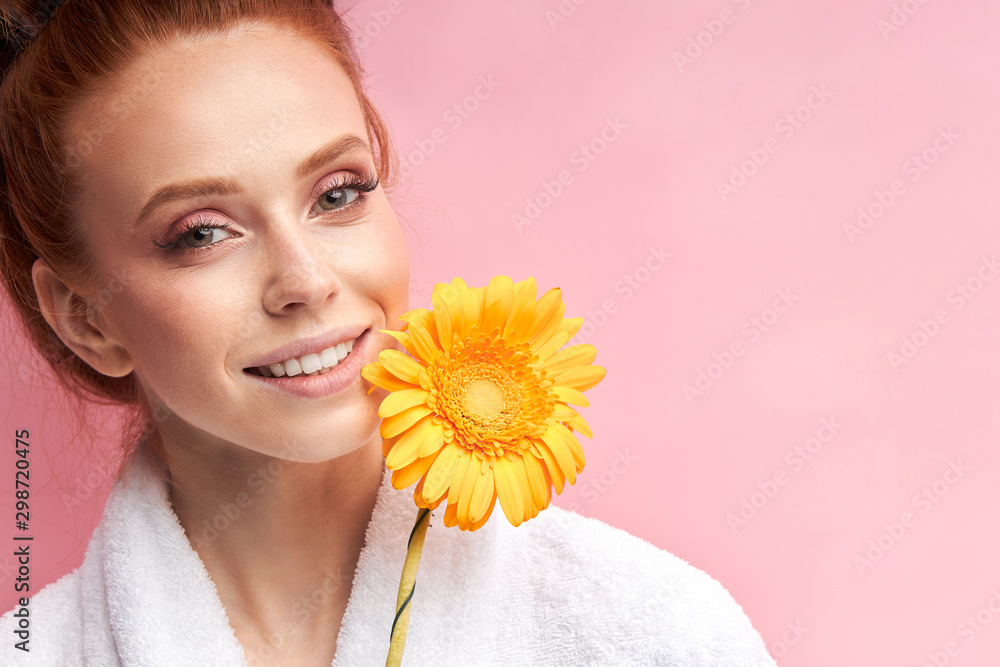 Smiling caucasian woman wearing white bathrobe, curlers on hair after shower, holding daisy flower. Natural beauty. Pink background Smiling caucasian woman wearing white bathrobe, curlers on hair after shower, holding daisy flower. Natural beauty. Pink background