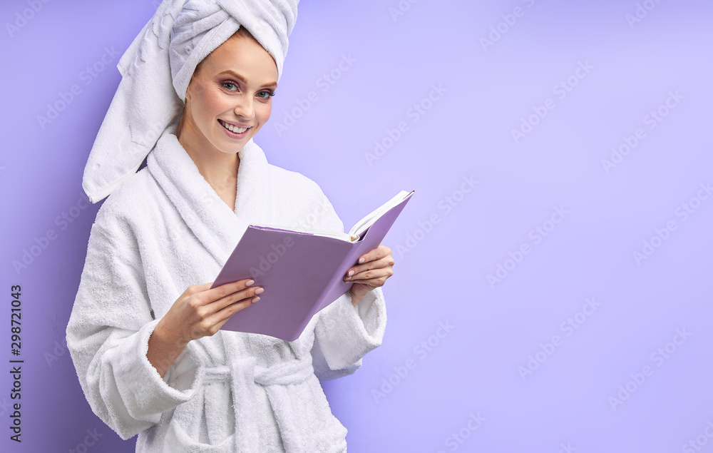Beautiful woman in bathrobe and towel after shower reading book isolated over purple background. Emotions, reading Beautiful woman in bathrobe and towel after shower reading book isolated over purple background. Emotions, reading