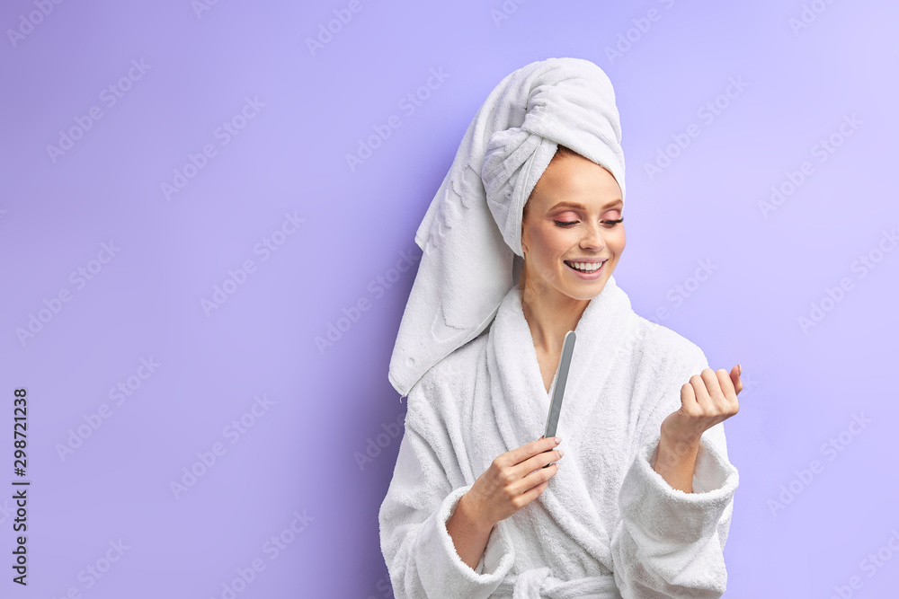 beautiful woman in bathrobe and towel on head cuts nails with nailfile, look at nails. Isolated over purple background beautiful woman in bathrobe and towel on head cuts nails with nailfile, look at nails. Isolated over purple background