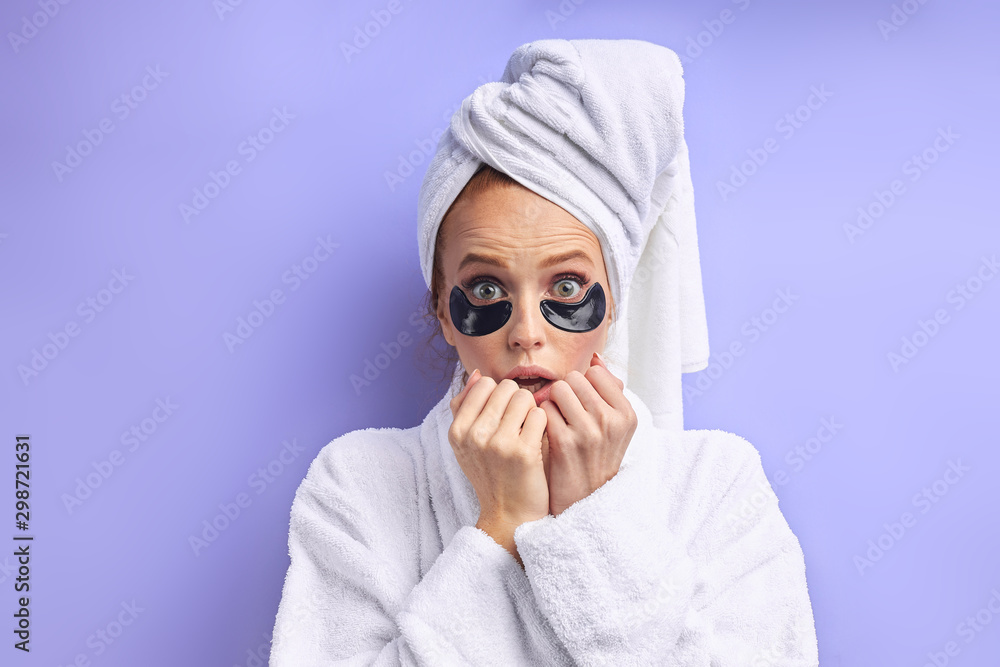 Caucasian female in bathrobe and towel posing with black patches under eyes. Surprised girl isolated over purple background Caucasian female in bathrobe and towel posing with black patches under eyes. Surprised girl isolated over purple background