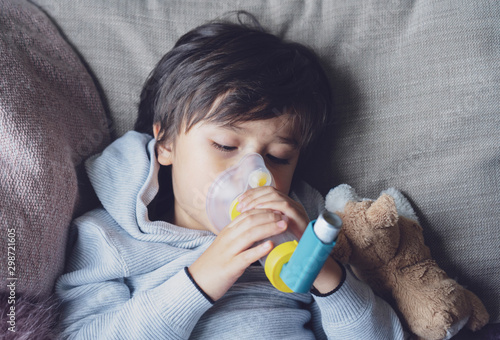 Poor boy tired from chest coughing holding inhaler mask, Child closing his eyes while using the volumtic for breathing treatment,Tried Kid having asthma allergy using the asthma inhaler photo