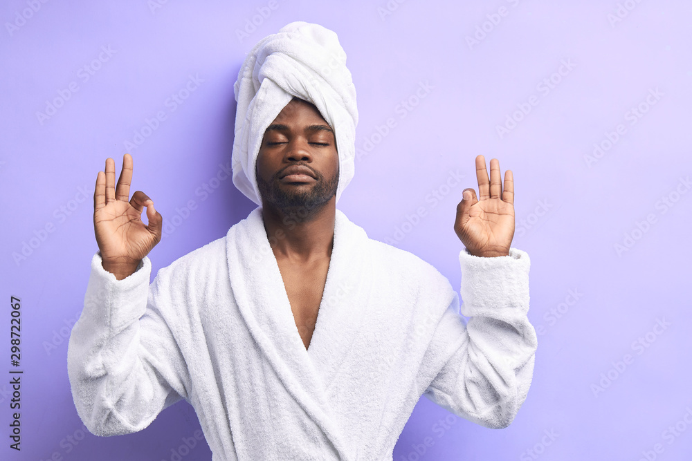 Concentrated african man in lotus position cropped. Meditating with closed eyes. Male in bathrobe and towel isolated over purple background Concentrated african man in lotus position cropped. Meditating with closed eyes. Male in bathrobe and towel isolated over purple background