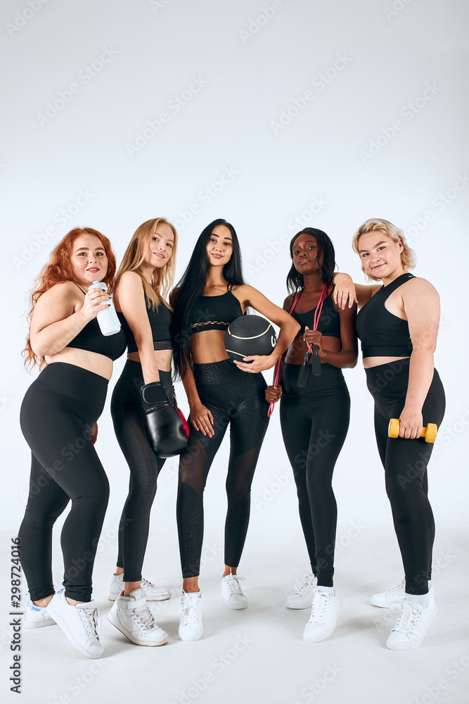 Fototapeta premium Five smiling multiethnic women wearing sportive leggins and topics holding fitness ball, boxing gloves and other things isolated over white background