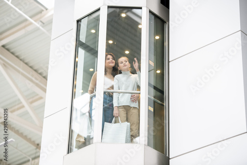 Young mother and daughter discussing something while moving upwards in elevator