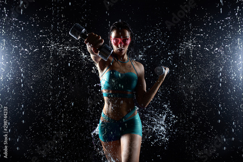 Beautiful young girl in sportswear poses with dumbbells in aqua studio. Drops of water spread about her fitness body. The perfect figure on the background of water splashes