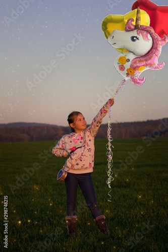 A ten year old girl stands in a field holding helium balloons - a red and gold heart and a unicorn.Little girl in sweatshirt with handbag standing on field and holding balloons. Fireflies fly in the g photo