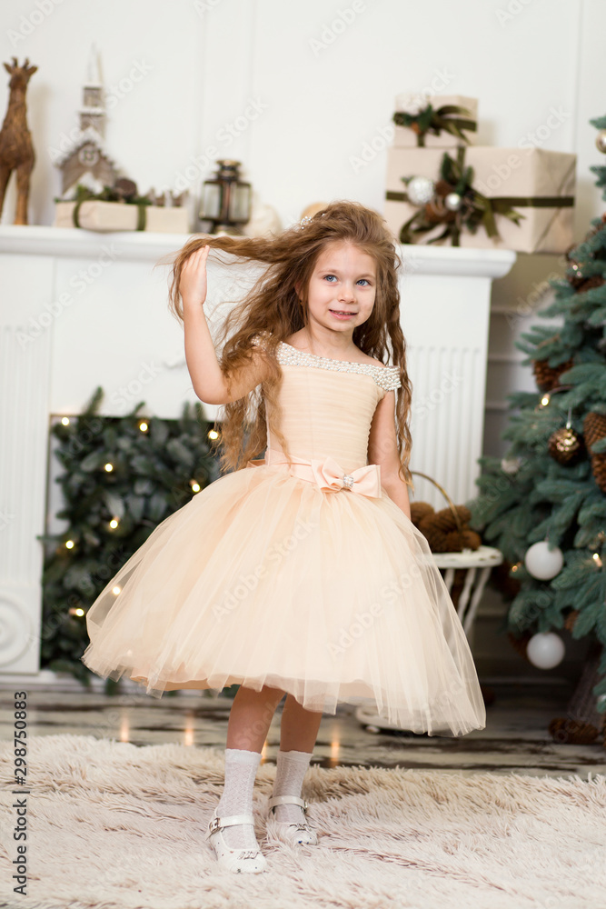 Little cute girl princess in a chic dress laughs and poses near the Christmas tree Little cute girl princess in a chic dress laughs and poses near the Christmas tree