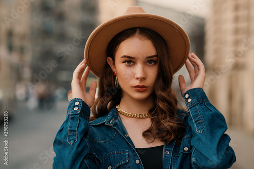 Outdoor close up fashion portrait of young elegant lady wearing beige fedora hat, trendy chain necklace, blue denim shirt, posing in street of European city. Copy, empty space for text