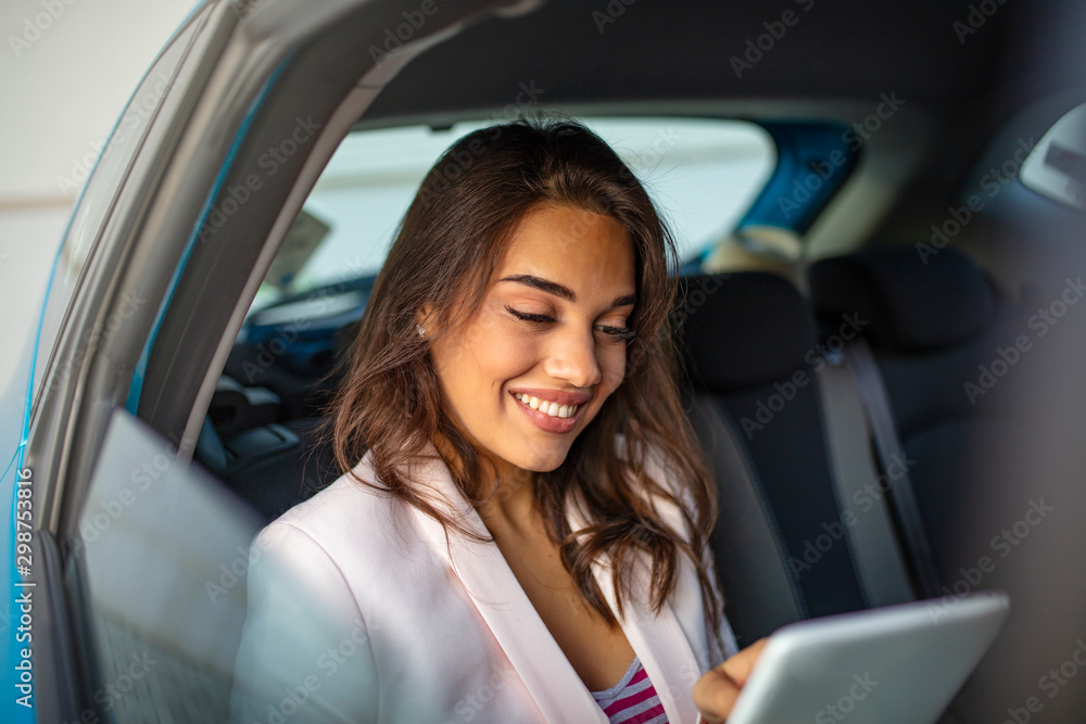 Young, beautiful woman sitting in the back seat of the car with a tablet in hand and drinking coffee. Close up portrait of a young business woman using digital tablet in the back seat of the car.