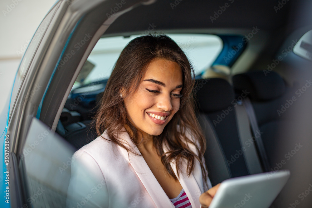 Young, beautiful woman sitting in the back seat of the car with a tablet in hand and drinking coffee. Close up portrait of a young business woman using digital tablet in the back seat of the car. Young, beautiful woman sitting in the back seat of the car with a tablet in hand and drinking coffee. Close up portrait of a young business woman using digital tablet in the back seat of the car.