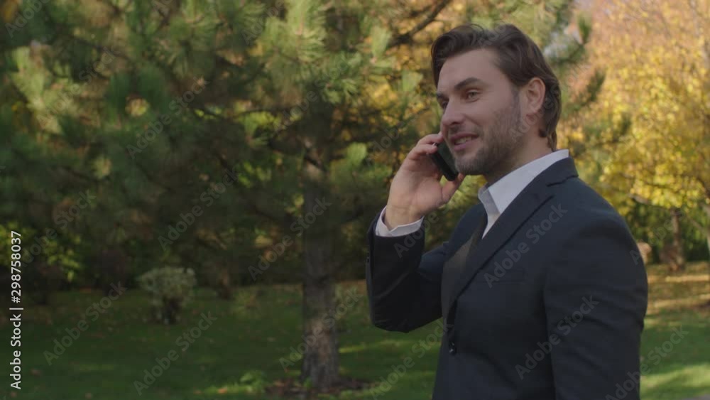 A handsome young guy of European appearance with a slight unshaven is having a pleasant conversation on the phone on the background of a green park. Smiling A handsome young guy of European appearance with a slight unshaven is having a pleasant conversation on the phone on the background of a green park. Smiling