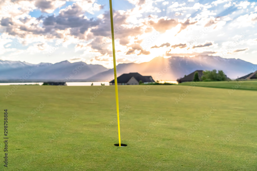 Obraz premium Yellow flagstick and hole at a golf course against lake mountain and cloudy sky