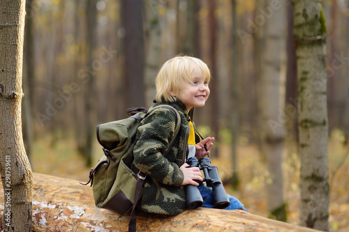 Little boy scout with binoculars during hiking in autumn forest. Child is sitting on large fallen tree and looking through a binoculars.