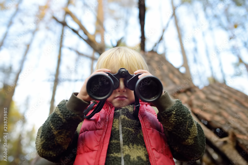Little boy scout is looking through binoculars during hiking in autumn forest. Behind the child is teepee hut. Little boy scout is looking through binoculars during hiking in autumn forest. Behind the child is teepee hut.