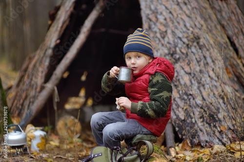 Little boy scout during hike in forest on autumn day. Child is cooking tea with help tourist gas burner and eating sandwich. Behind the child is teepee hut.