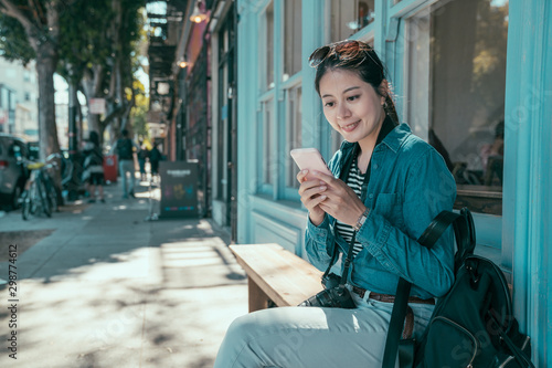Stylish asian girl backpacker using apps on cellphone while sitting on bench on citywalk outside store during summer vacation. Female blogger reading article on mobile phone relax on street.