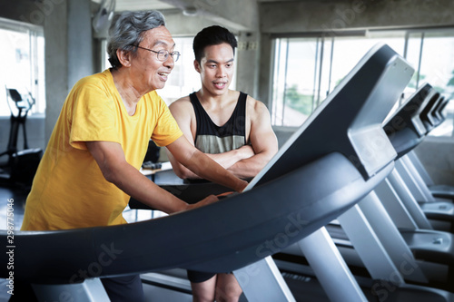 Elderly men are walking exercise on machine treadmill at fitness gym club with a personal trainer to take care of counseling.