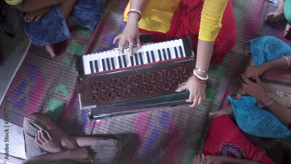 Married Indian teacher teaches music with harmonium at rural classroom with students gathered