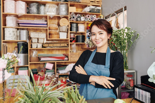 Portrait of young positive florist in blue apron standing at counter with her arms folded and smiling at camera