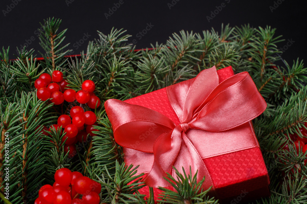 New Year Christmas Xmas 2020 holiday celebration red present gift box with satin pink bow, immersed in the needles of a Christmas tree decorated with red berries. Dark festive composition. New Year Christmas Xmas 2020 holiday celebration red present gift box with satin pink bow, immersed in the needles of a Christmas tree decorated with red berries. Dark festive composition.