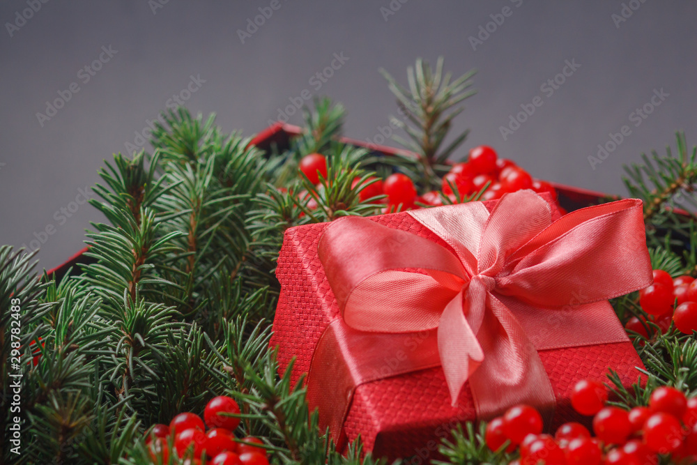 New Year Christmas Xmas 2020 holiday celebration red present gift box with satin pink bow, immersed in the needles of a Christmas tree decorated with red berries. Dark festive composition. New Year Christmas Xmas 2020 holiday celebration red present gift box with satin pink bow, immersed in the needles of a Christmas tree decorated with red berries. Dark festive composition.