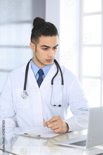 Arab doctor man using laptop computer while filling up medication history records form at the glass desk in medical office or clinic. Medicine and healthcare concept