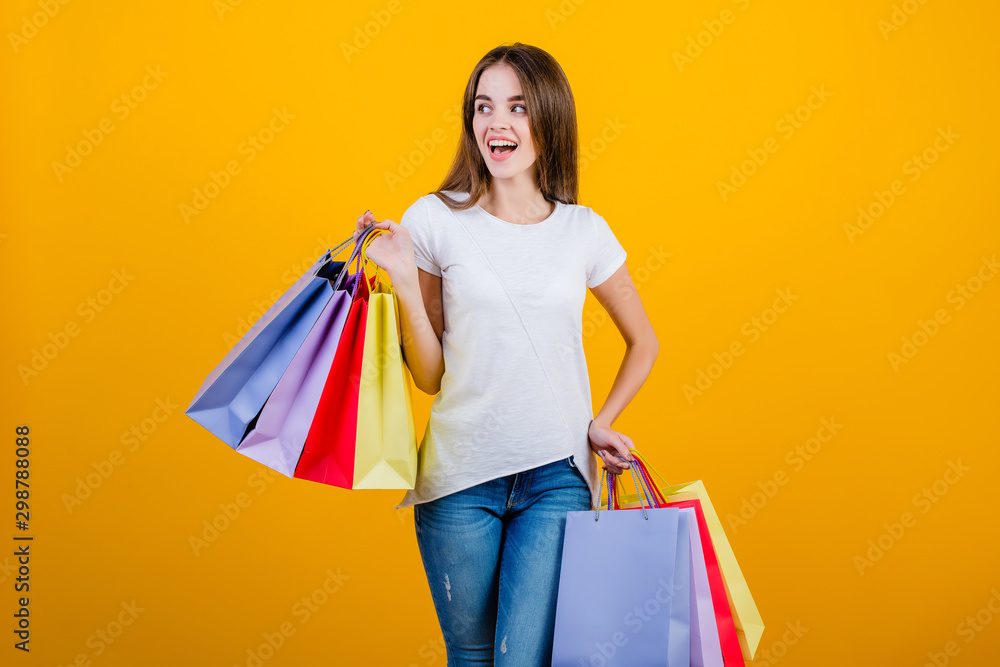 happy smiling beautiful brunette woman with colorful paper shopping bags wearing jeans and shirt isolated over yellow