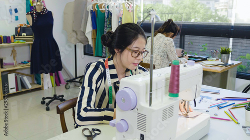 beautiful smiling woman fashion designer using sewing machine sitting at desk in modern studio. hard working lady dressmaker colleague in back drawing on sketch paper. two teamwork partner girls.