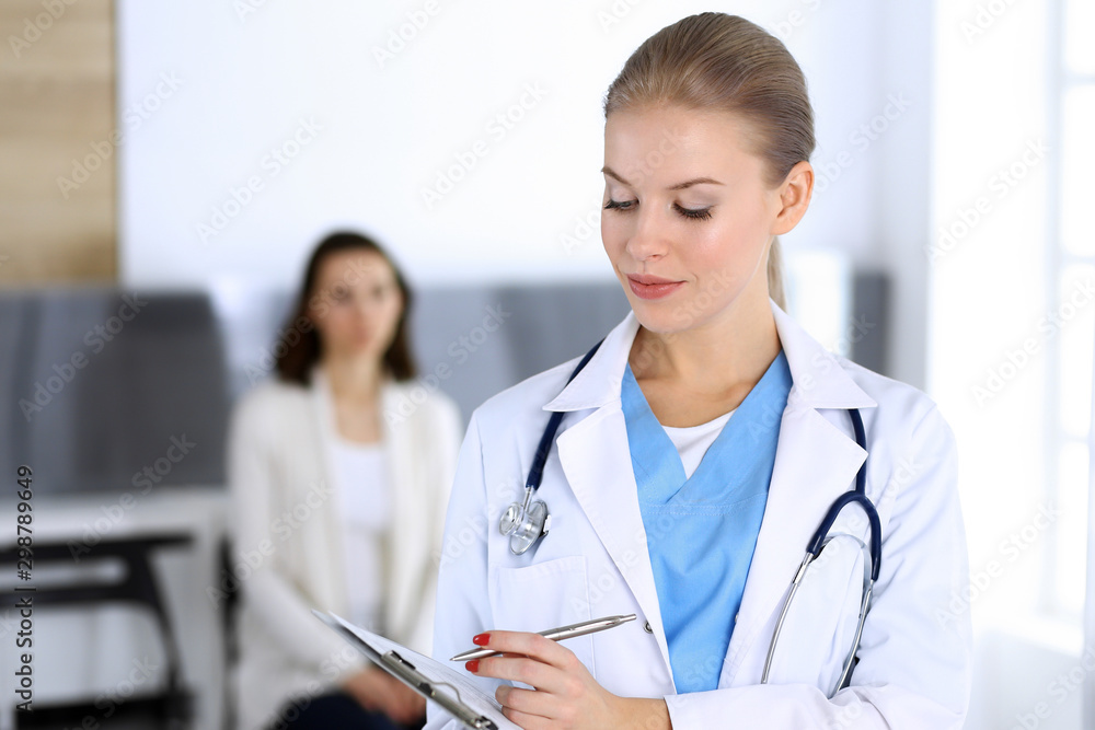 Doctor woman filling up medication history record while standing in emergency hospital office with patient in queue at background. Physician at work, portrait shoot. Medicine and health care concept Doctor woman filling up medication history record while standing in emergency hospital office with patient in queue at background. Physician at work, portrait shoot. Medicine and health care concept