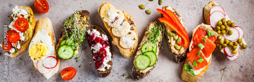 Open toasts with different toppings on gray-brown background, top view. Flat lay of crostini with banana, pate, avocado, salmon, egg, cheese and berries.