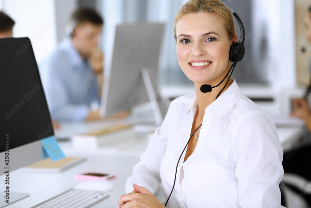 Blonde business woman using headset for communication and consulting people at customer service office. Call center. Group of operators at work at the background Blonde business woman using headset for communication and consulting people at customer service office. Call center. Group of operators at work at the background