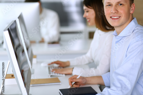 Cheerful smiling businessman headshot at work in modern office. Unknown casual dressed entrepreneur using pc computer while sitting with diverse colleagues at the background. Multi ethnic working