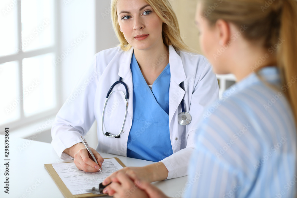 Woman doctor and patient sitting and talking at medical examination in hospital or clinic. Blonde therapist cheerfully smiling. Medicine and healthcare concept Woman doctor and patient sitting and talking at medical examination in hospital or clinic. Blonde therapist cheerfully smiling. Medicine and healthcare concept
