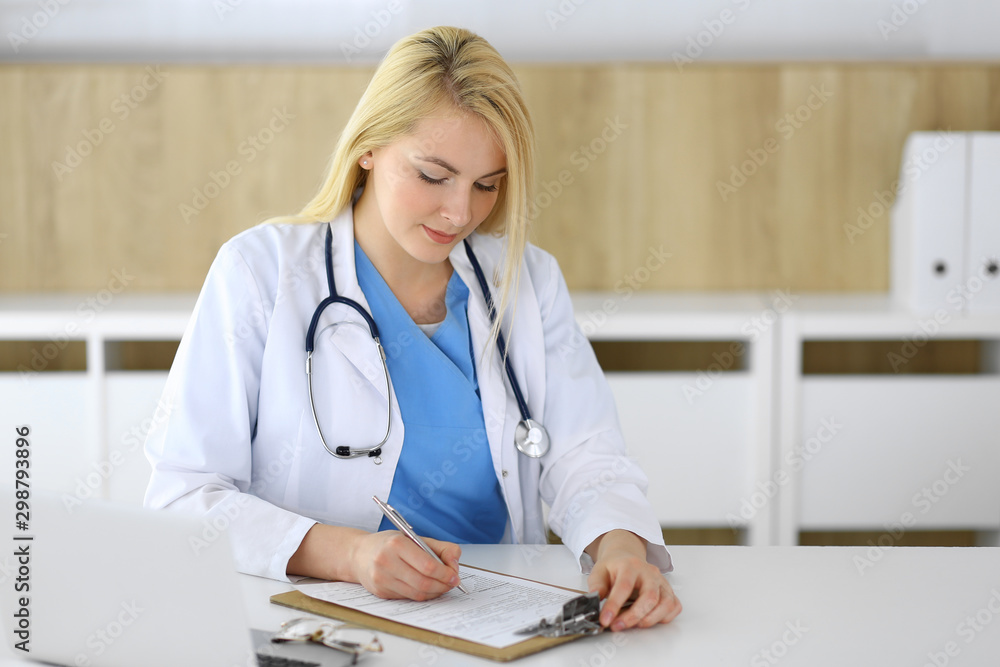 Doctor woman at work while sitting at the desk in hospital or clinic. Blonde cheerful physician filling up medical records form. Data and best service in medicine and healthcare Doctor woman at work while sitting at the desk in hospital or clinic. Blonde cheerful physician filling up medical records form. Data and best service in medicine and healthcare