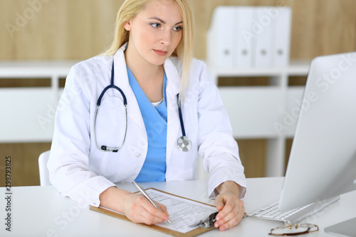 Doctor woman at work while sitting at the desk in hospital or clinic. Blonde cheerful physician filling up medical records form. Data and best service in medicine and healthcare