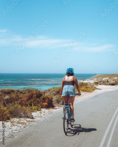 Woman riding a bicycle along a bike path with the beautiful blue ocean as the backdrop, at Rottnest Island in Perth, Western Australia.