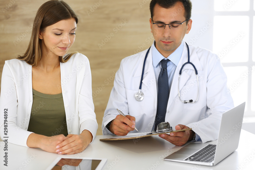 Doctor and patient discussing the results of a physical examination while sitting at a desk in a clinic. A male doctor using a clipboard to fill out a medical history of a young woman's medication Doctor and patient discussing the results of a physical examination while sitting at a desk in a clinic. A male doctor using a clipboard to fill out a medical history of a young woman's medication