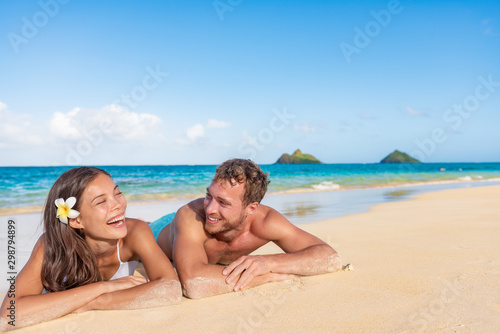 Happy interracial couple beach vacation summer fun relaxing laughing together. Hawaii travel holidays. Young love on romantic vacations suntan lying down on sand. Asian girl, Caucasian man.