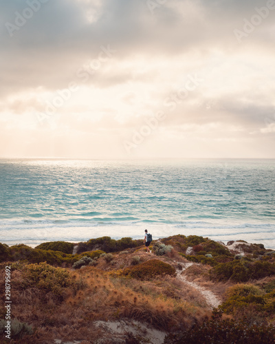 Lone man with with professional camera backpack, walking through the winding sand path through the sand dunes covered in greenery, walking towards the beautiful ocean and bright sky with rays shining. photo