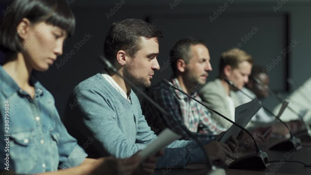 Tracking backlit side view shot of multiethnic group of businesspeople discussing contract details at business conference. Focus on young Middle Eastern businessman speaking into microphone in meeting Tracking backlit side view shot of multiethnic group of businesspeople discussing contract details at business conference. Focus on young Middle Eastern businessman speaking into microphone in meeting