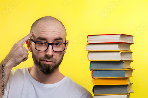 Concept of education. A bald, bearded man with glasses points a finger at his head. Nearby is a stack of books. Yellow background