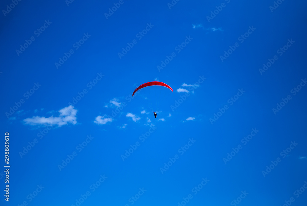 The red paraglider flies high above, with only blue skies and a few small clouds behind. The red paraglider flies high above, with only blue skies and a few small clouds behind.