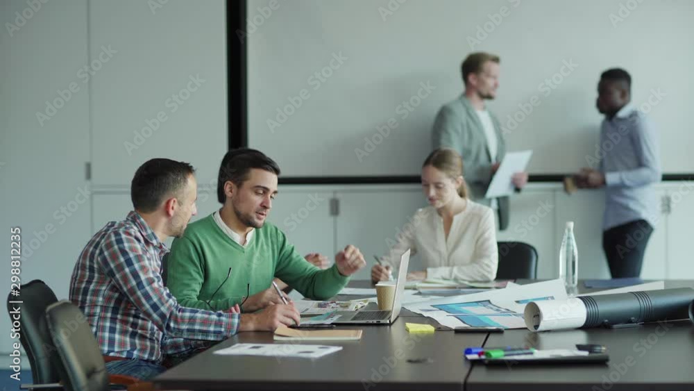 Group of multiethnic business people working in office. Two men discussing ideas at desk, female colleagues talking and writing in notebook nearby. Two businessmen discussing in background Group of multiethnic business people working in office. Two men discussing ideas at desk, female colleagues talking and writing in notebook nearby. Two businessmen discussing in background