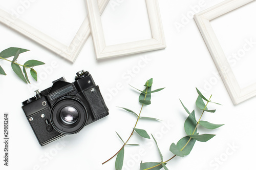 White photo frame, old retro photo camera and green eucalyptus leaves on white background. Flat lay top view copy space. Stylish minimal composition, artwork mockup, picture frame, home decoration photo