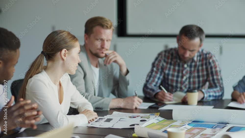 Tracking shot of diverse business team discussing market analysis sitting at desk in meeting. Young woman holding document with charts and explaining statistics to colleagues Tracking shot of diverse business team discussing market analysis sitting at desk in meeting. Young woman holding document with charts and explaining statistics to colleagues
